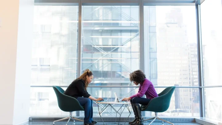 Two coworkers in a quietly tense conversation across a desk in an office